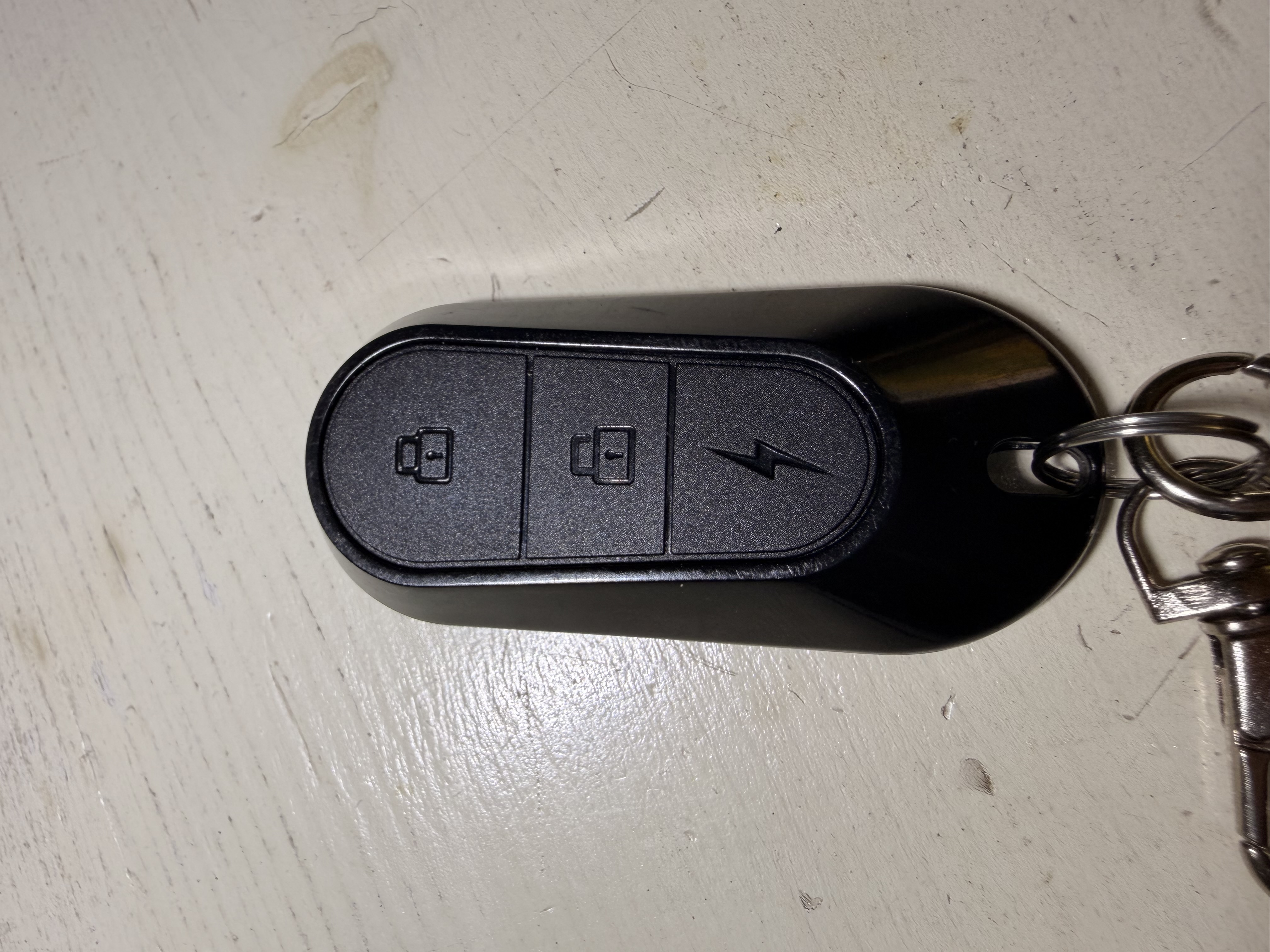 Telecomando frontale: 3 tasti — LOCK 🔒 (auto chiuso), UNLOCK 🔓 (auto aperto), PANIC ⚡ (fulmine = sirena/SOS)
