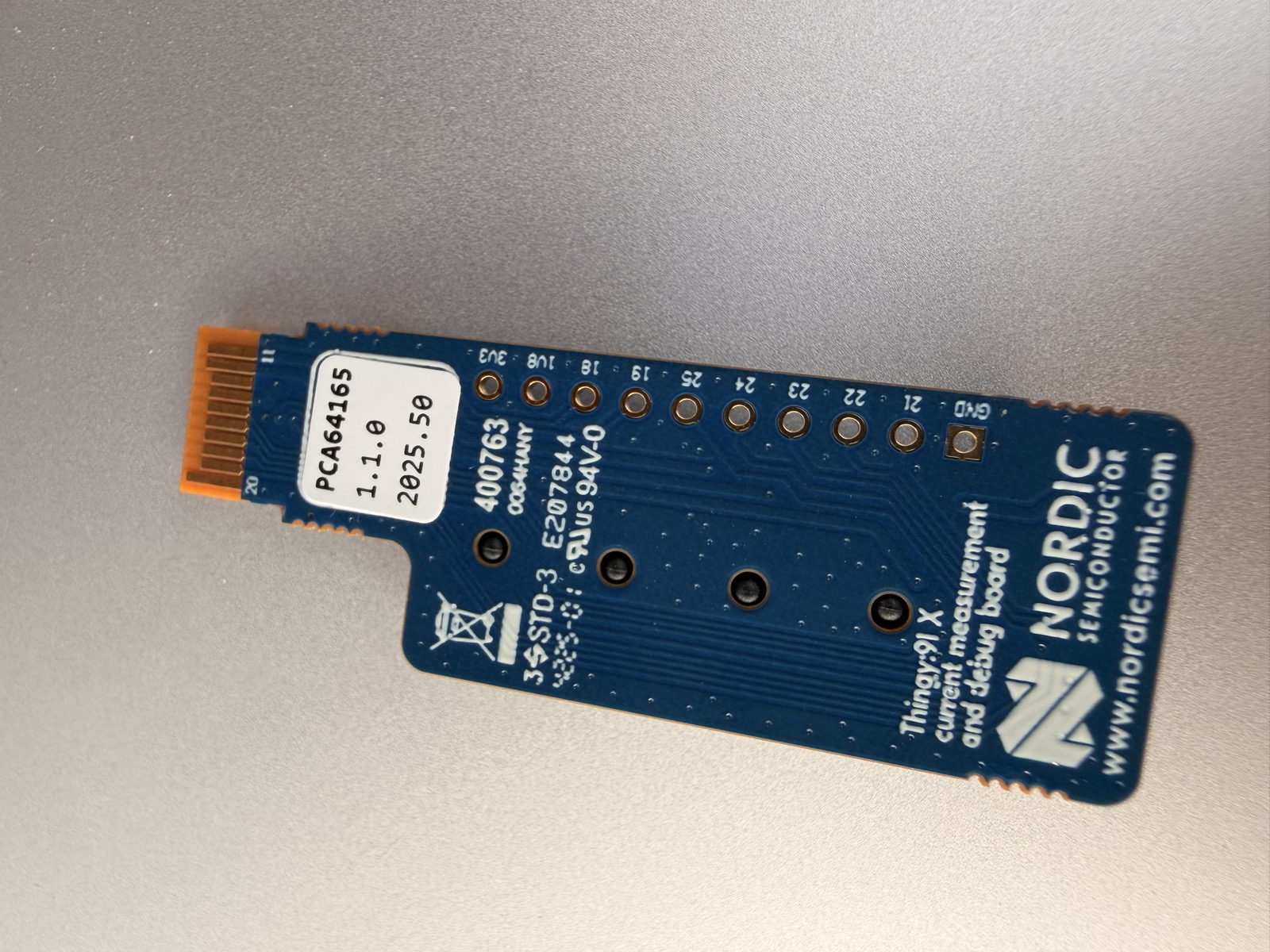 Thingy:91 X current measurement & debug board PCA64165 — lato saldature con logo Nordic, etichetta PCA64165 v1.1.0 2025.50, FPC arancione 20-pin